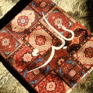 Könnte beinhalten: Eine dekorative Box mit einem lebendigen, gemusterten Design, das an einen Orientteppich erinnert. Die Box weist ein sich wiederholendes Muster aus roten, blauen und cremefarbenen Blumenmotiven auf. Weiße Schrift ist auf die Box gelegt. Die Box steht auf einer strukturierten, beigefarbenen Oberfläche.