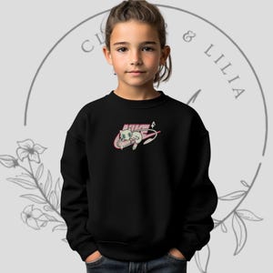 Könnte beinhalten: Ein schwarzes Sweatshirt mit einem Cartoon-Charakter-Design in Rosa und Weiß. Das Design befindet sich in einem rosa Swoosh, mit dem Wort "NIKE" darüber. Das Sweatshirt wird von einer jungen Person getragen.