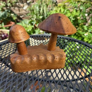 May include: A wooden mushroom sculpture featuring two mushrooms of different sizes. The mushrooms are brown with a smooth finish and sit on a carved wooden base. The sculpture is displayed on a black metal table outdoors.