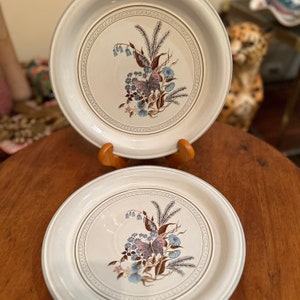 May include: Two white ceramic plates with a brown, blue, and purple floral design featuring a butterfly. The plates have a gold rim and are on a wooden stand.
