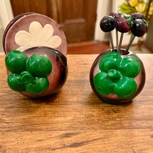 May include: A set of two ceramic napkin holders and a toothpick holder, all shaped like mangosteens. The napkin holders have a white flower design and the toothpick holder has a green flower design with four black toothpicks with purple berries on top.