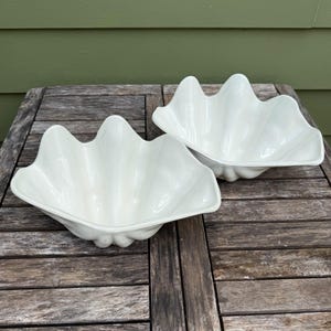 May include: Two white, shell-shaped decorative bowls. The bowls have a scalloped edge and a glossy finish. They are placed on a weathered wooden table. The bowls are a decorative item, suitable for coastal or nautical-themed decor.