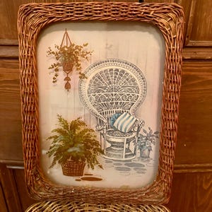 May include: A rectangular wicker picture frame with a vintage print of a peacock chair, potted plants, and a hanging basket. The print has a soft, muted color palette of blue, green, and beige. The frame is made of woven brown wicker.