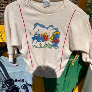 May include: A white long-sleeved sweatshirt with red trim on the sleeves. The front of the sweatshirt features a cartoon image of Smurfette and a Smurf holding a gift box. The text "Cheroy M" is visible on the tag.
