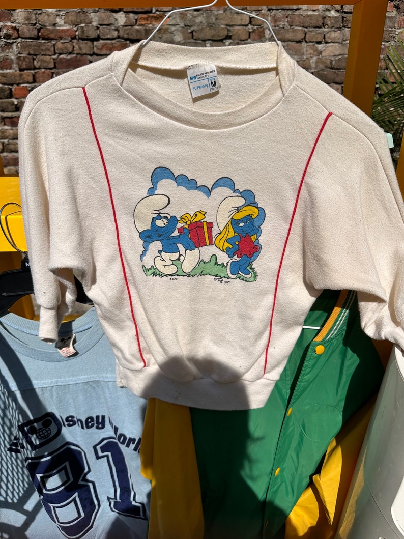 May include: A white long-sleeved sweatshirt with red trim on the sleeves. The front of the sweatshirt features a cartoon image of Smurfette and a Smurf holding a gift box. The text "Cheroy M" is visible on the tag.