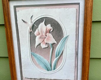 May include: Framed floral art print featuring a pink lily and a hummingbird. The artwork is set within a light pink and gray oval, with green leaves and a brown wooden frame. The print is on a textured paper.