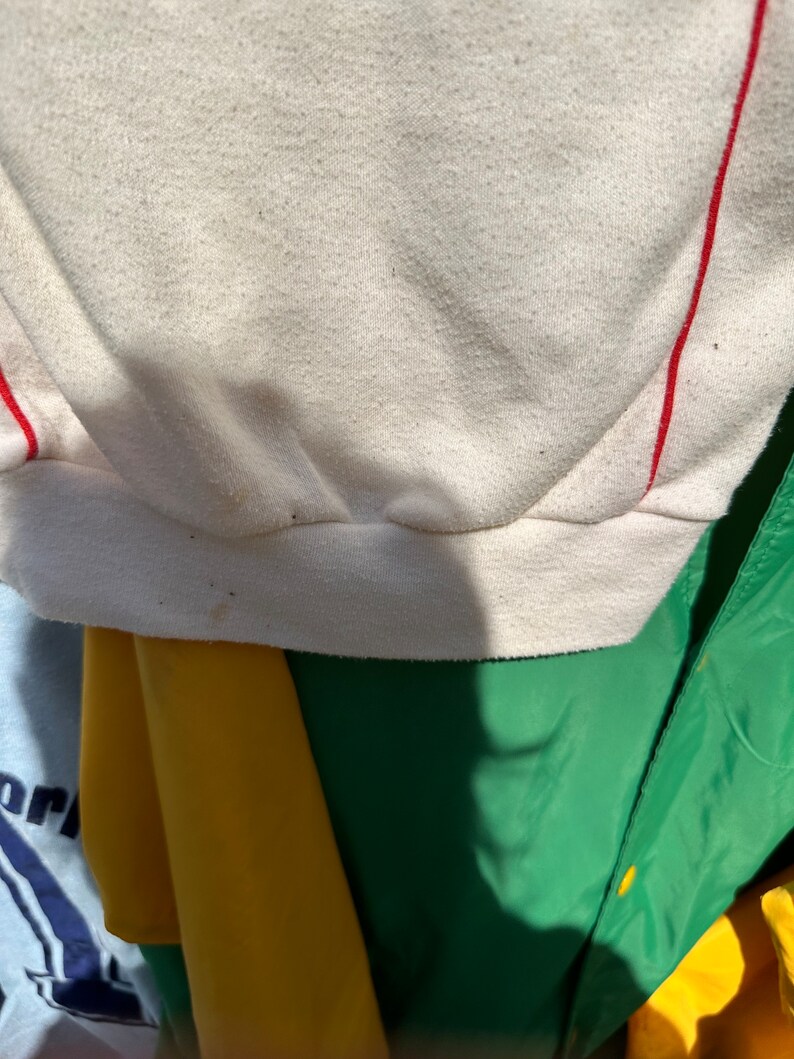 May include: Close-up of the bottom edge of a white sweatshirt with red stripes. The fabric is textured and has a slight sheen.