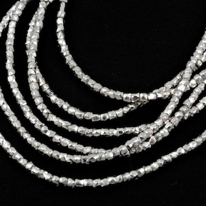 May include: A multi-strand necklace featuring small, irregularly shaped silver beads. The beads are strung together, creating a layered effect. The necklace is displayed against a black background, highlighting the silver color.