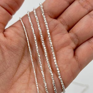 May include: Close-up of several delicate silver necklaces with faceted beads. The necklaces vary in thickness and are displayed against a neutral background. The silver chains have a reflective quality, catching the light.