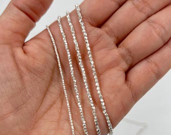 Puede incluir: Primer plano de varios delicados collares de plata con cuentas facetadas. Los collares varían en grosor y se muestran sobre un fondo neutro. Las cadenas de plata tienen una calidad reflectante, captando la luz.