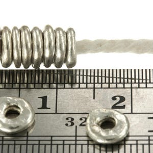 May include: Eight small, round, silver-colored metal beads with a textured, organic finish are strung on a white cord. Two additional loose beads are placed on a metal ruler, showing a diameter of approximately 0.3 inches for each bead.