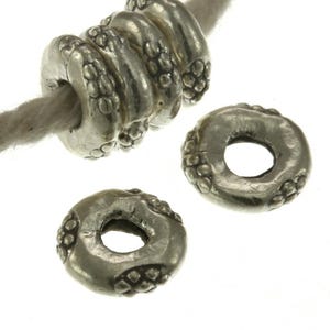 May include: Three silver-toned metal beads with a circular, donut-like shape and a textured, dotted pattern along the outer edges. One bead is threaded onto a light-colored cord, while two identical beads are positioned separately below it.