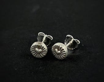 May include: A pair of small, silver stud earrings. Each earring features a circular design with a heart in the center, surrounded by radiating lines. The earrings have a post and butterfly backing.