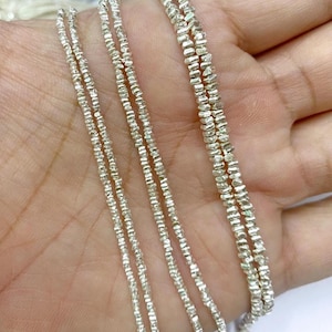 Può includere: Primo piano di diversi fili di delicate collane di perline color argento. Le perline hanno una forma sfaccettata e irregolare, che cattura la luce e crea un effetto scintillante. Le collane sono esposte su uno sfondo neutro, evidenziando il loro design intricato.