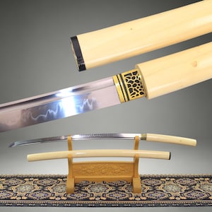 May include: A Japanese katana sword with a curved steel blade featuring a wavy hamon line. The handle and scabbard are made of smooth, cream-colored material. A gold-colored metal collar with a black honeycomb pattern sits near the blade base.
