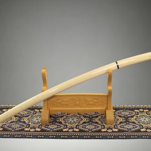 May include: A long, cylindrical, cream-colored sword scabbard with a smooth, polished finish and a single thin black band near the top, resting diagonally on a light-colored wooden stand featuring carved relief details on its base.