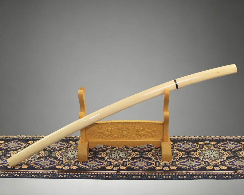 May include: A long, cylindrical, cream-colored sword scabbard with a smooth, polished finish and a single thin black band near the top, resting diagonally on a light-colored wooden stand featuring carved relief details on its base.