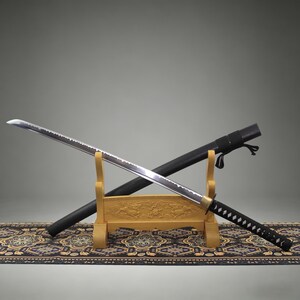 May include: A katana sword with a silver steel blade, a black wrapped handle, and a gold-colored guard. The sword rests on a light-colored wooden stand featuring carved details, positioned in front of a black scabbard with a black cord attachment.