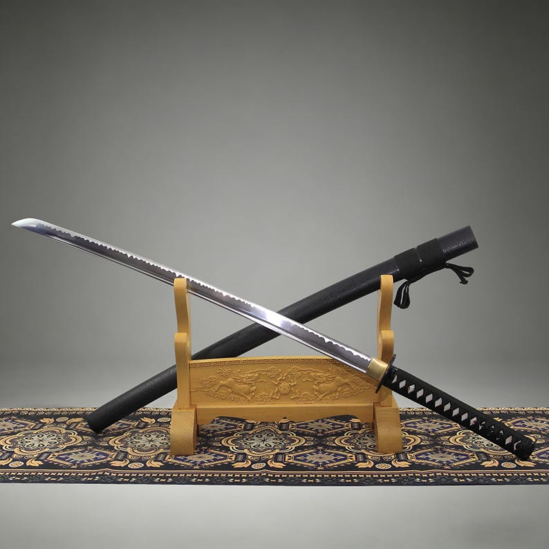 May include: A katana sword with a silver steel blade, a black wrapped handle, and a gold-colored guard. The sword rests on a light-colored wooden stand featuring carved details, positioned in front of a black scabbard with a black cord attachment.