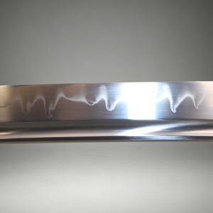 May include: A close-up view of a polished steel sword blade featuring a distinct, wavy hamon line pattern running along the edge. The metal surface is reflective, showing a smooth, metallic texture with a sharp, curved cutting edge.
