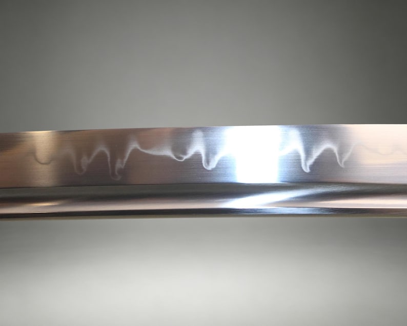 May include: A close-up view of a polished steel sword blade featuring a distinct, wavy hamon line pattern running along the edge. The metal surface is reflective, showing a smooth, metallic texture with a sharp, curved cutting edge.