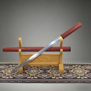 May include: A straight sword with a polished steel blade featuring a wavy hamon line, a reddish-brown wooden handle with two small brass pins, and a matching reddish-brown wooden scabbard resting on a light-colored carved wooden display stand.