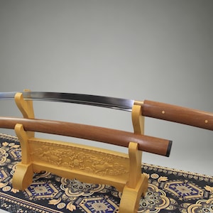 May include: A curved steel katana blade with a polished finish and a wooden handle, resting on a light-colored wooden display stand. The matching wooden scabbard is positioned horizontally on the stand below the blade, featuring carved details.