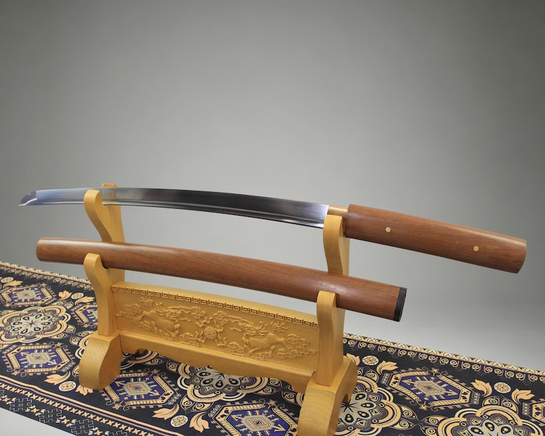 May include: A curved steel katana blade with a polished finish and a wooden handle, resting on a light-colored wooden display stand. The matching wooden scabbard is positioned horizontally on the stand below the blade, featuring carved details.