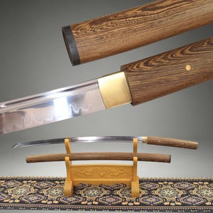 Può includere: Una spada katana giapponese con una lama in acciaio lucido caratterizzata da una linea di tempra ondulata visibile, un manico in legno e un fodero in legno abbinato. La spada poggia su un supporto in legno chiaro con dettagli intagliati.