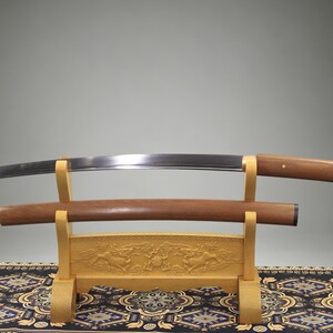 May include: A curved steel sword blade with a wooden handle and a separate wooden scabbard resting on a light-colored wooden display stand. The stand features intricate carved details and is supported by two vertical uprights with curved supports.