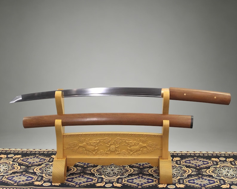 May include: A curved steel sword blade with a wooden handle and a separate wooden scabbard resting on a light-colored wooden display stand. The stand features intricate carved details and is supported by two vertical uprights with curved supports.