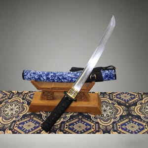 May include: A Japanese katana sword with a curved steel blade, a black wrapped handle, and a gold-toned metal guard. The scabbard is blue and white with a floral pattern and black cord. The sword rests on a brown wooden stand.
