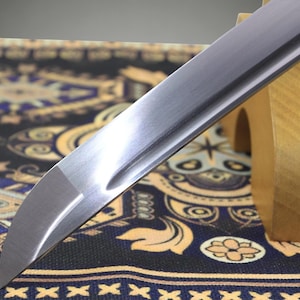 May include: A silver-colored metal sword blade with a curved tip and a central groove running along the length of the steel. The blade features a polished finish and a sharp edge, resting at an angle against a light-colored wooden support stand.