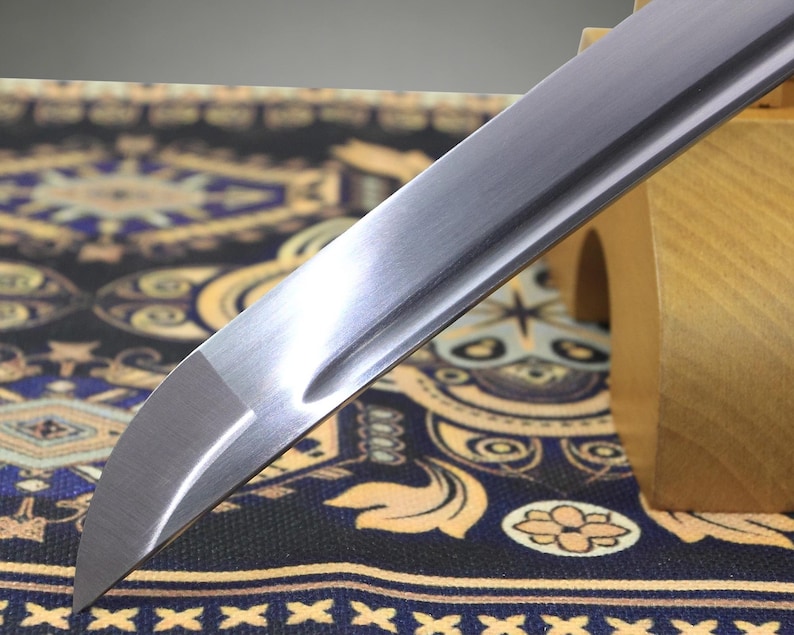 May include: A silver-colored metal sword blade with a curved tip and a central groove running along the length of the steel. The blade features a polished finish and a sharp edge, resting at an angle against a light-colored wooden support stand.