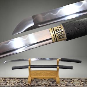 May include: A katana sword with a curved steel blade featuring a visible wavy temper line, a gold-colored metal collar with a cutout pattern, and a black textured handle. The sword is displayed on a light-colored wooden stand with a black scabbard.