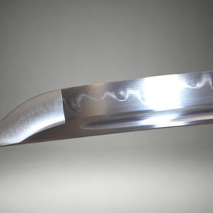 May include: A close-up view of a silver-colored steel sword blade featuring a curved tip and a distinct wavy hamon line running along the edge. The metal surface has a polished finish with a visible groove extending along the length of the blade.