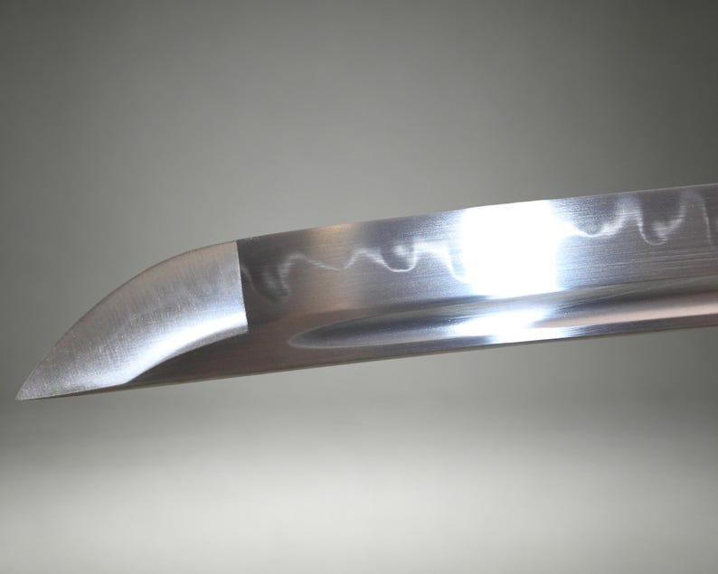 May include: A close-up view of a silver-colored steel sword blade featuring a curved tip and a distinct wavy hamon line running along the edge. The metal surface has a polished finish with a visible groove extending along the length of the blade.