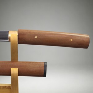 May include: A sword with a polished metal blade and a smooth brown wooden handle featuring two small circular brass inlays. The sword rests on a light-colored wooden stand, with a matching wooden scabbard positioned directly below the blade.