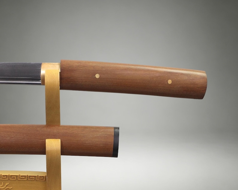 May include: A sword with a polished metal blade and a smooth brown wooden handle featuring two small circular brass inlays. The sword rests on a light-colored wooden stand, with a matching wooden scabbard positioned directly below the blade.
