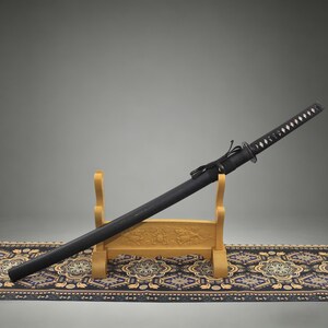 May include: A black katana sword with a wrapped handle featuring white diamond-shaped accents, housed in a matching black scabbard. The sword rests on a light-colored wooden display stand that has carved details on the front crossbar.