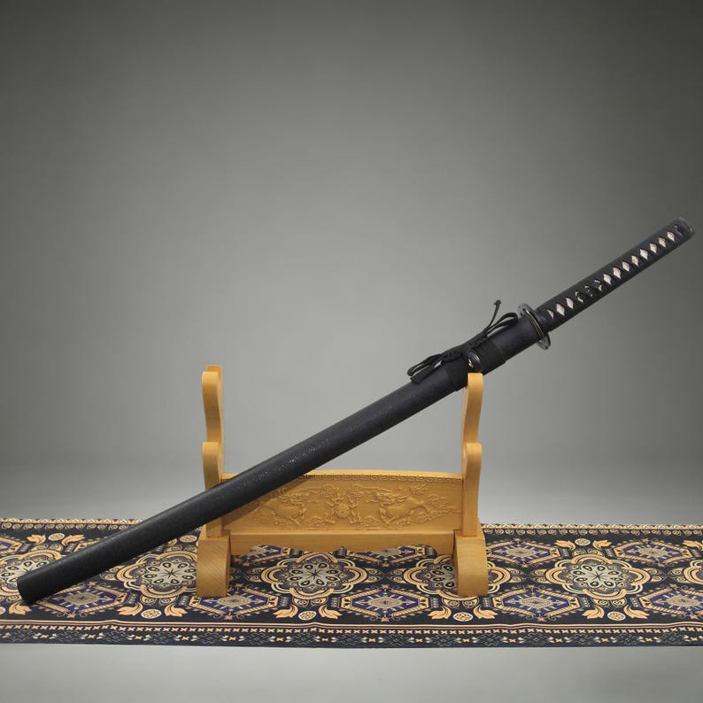 May include: A black katana sword with a wrapped handle featuring white diamond-shaped accents, housed in a matching black scabbard. The sword rests on a light-colored wooden display stand that has carved details on the front crossbar.