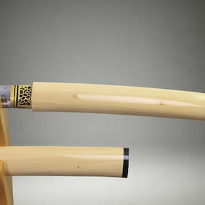 May include: A katana sword with a smooth, cream-colored handle and scabbard resting on a light wood stand. The sword features a metallic collar with a black and gold honeycomb pattern near the blade, which shows a visible wavy temper line.