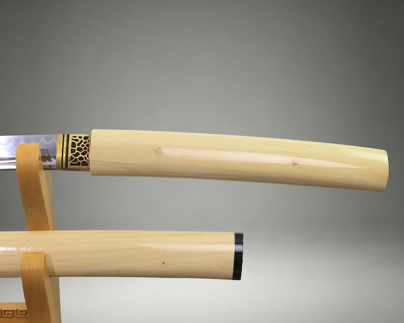 May include: A katana sword with a smooth, cream-colored handle and scabbard resting on a light wood stand. The sword features a metallic collar with a black and gold honeycomb pattern near the blade, which shows a visible wavy temper line.