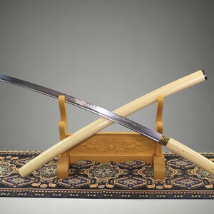 May include: A katana sword with a curved steel blade featuring a visible temper line and a light cream-colored wooden scabbard and handle. The sword rests on a carved wooden stand, with the scabbard positioned diagonally behind the blade.