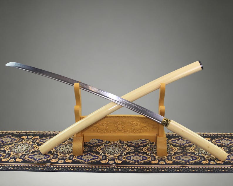 May include: A katana sword with a curved steel blade featuring a visible temper line and a light cream-colored wooden scabbard and handle. The sword rests on a carved wooden stand, with the scabbard positioned diagonally behind the blade.