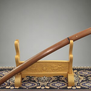 May include: A wooden sword scabbard with a smooth, polished brown finish, featuring two small circular brass inlays on the handle section and a thin black band near the center, resting on a light-colored carved wooden display stand.