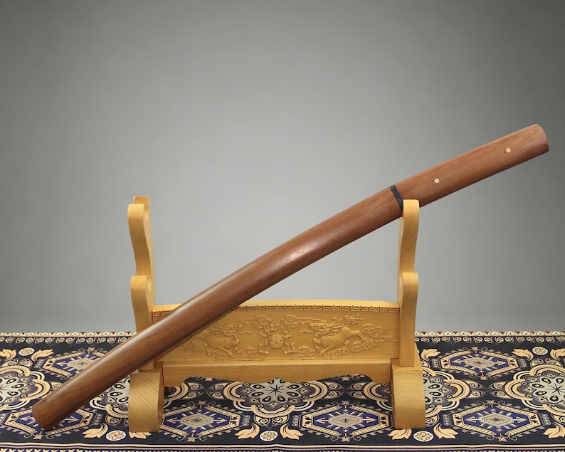 May include: A wooden sword scabbard with a smooth, polished brown finish, featuring two small circular brass inlays on the handle section and a thin black band near the center, resting on a light-colored carved wooden display stand.