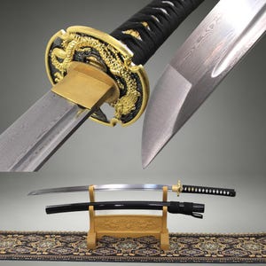 May include: A katana sword featuring a silver steel blade with a visible folded metal pattern, a black cord-wrapped handle, and a gold-coloured circular guard decorated with a dragon motif. The sword is displayed on a wooden stand with a black scabbard.