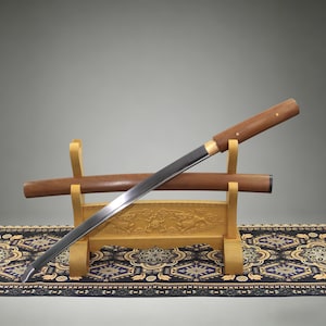 May include: A Japanese-style sword with a polished steel blade and a smooth wooden handle, resting on a light-colored wooden stand. The sword is positioned diagonally across its matching wooden scabbard, which is also supported by the stand.