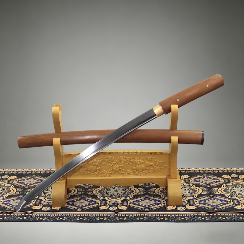 May include: A Japanese-style sword with a polished steel blade and a smooth wooden handle, resting on a light-colored wooden stand. The sword is positioned diagonally across its matching wooden scabbard, which is also supported by the stand.